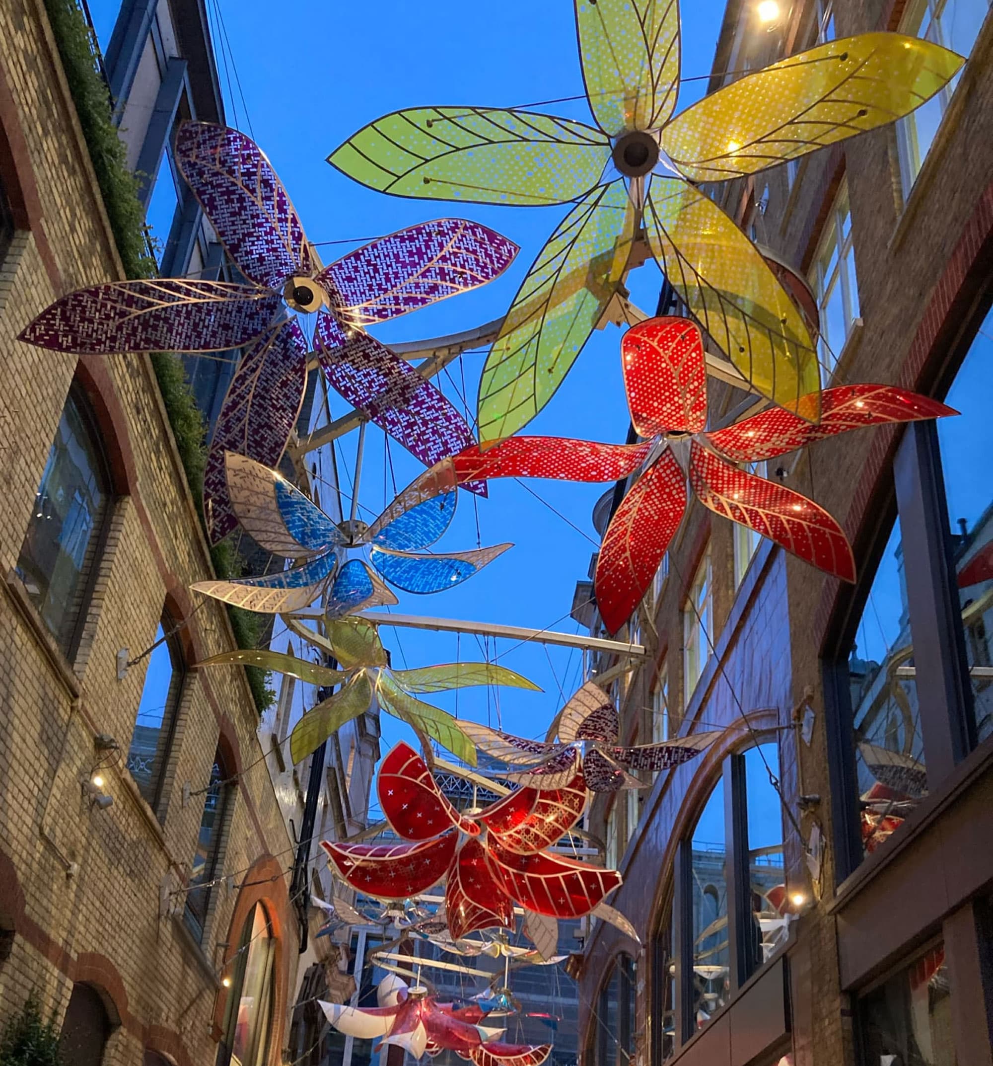 Bunte, leuchtende Blumeninstallationen aus Glas oder Kunststoff hängen zwischen Gebäuden in einer schmalen Gasse, beleuchtet am Abendhimmel.