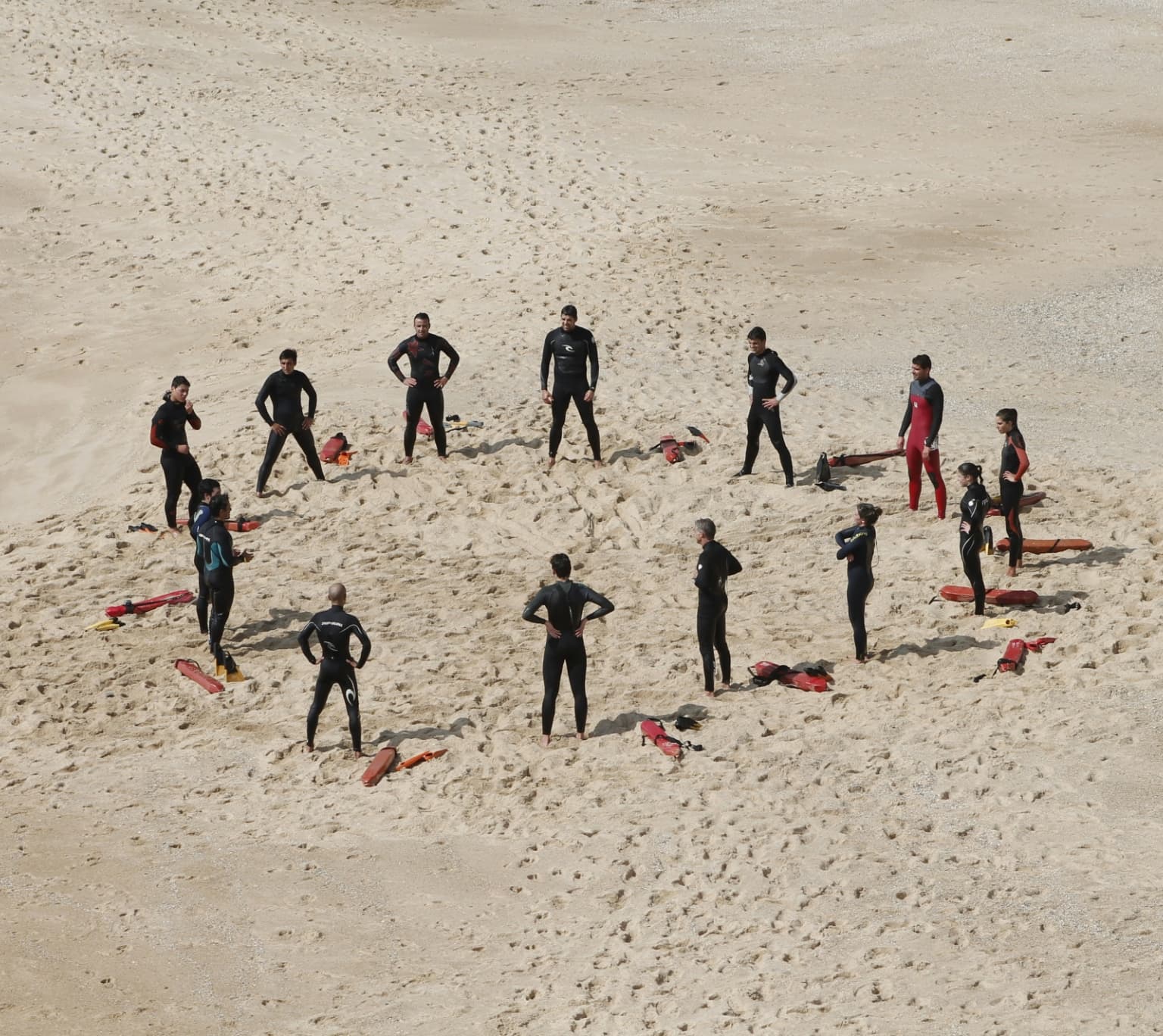 Gruppe von Menschen in Neoprenanzügen steht im Kreis auf einem Sandstrand und scheint sich auf eine Aktivität vorzubereiten, möglicherweise ein Surf- oder Rettungstraining.